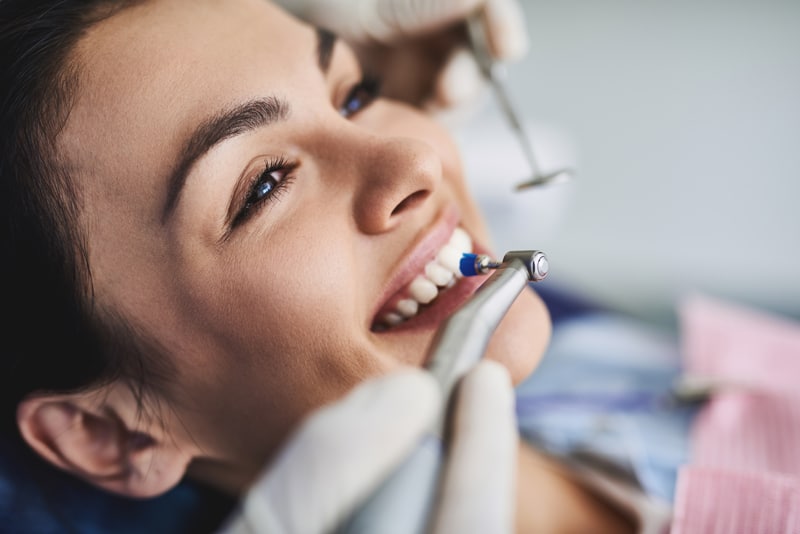 A woman smiles during a dental cleaning as a dentistâs gloved hands hold tools near her teeth. She wears a pink bib and looks relaxed.