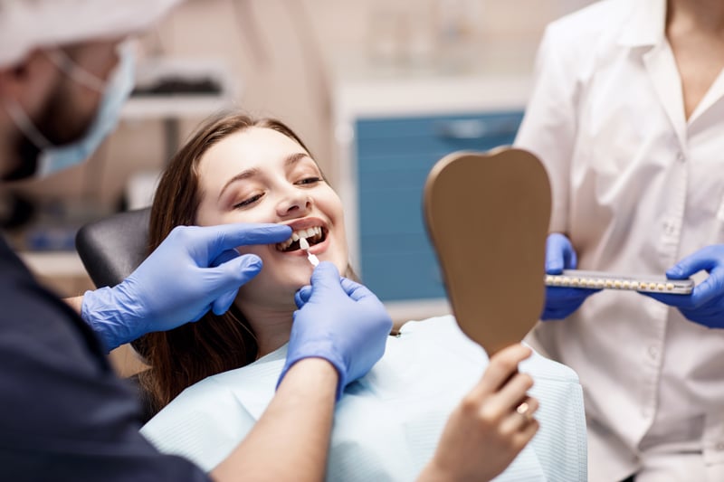 A woman smiles in a dental chair holding a tooth-shaped mirror as a dentist checks her teeth with a shade guide; assistant nearby.