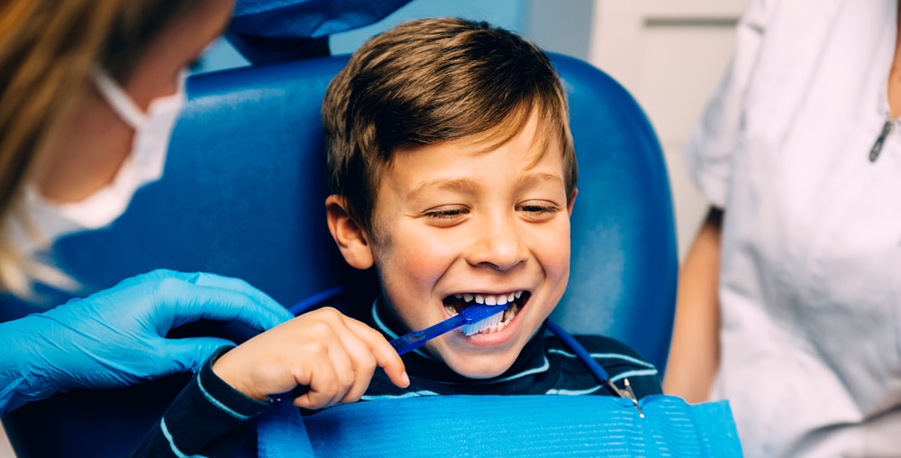 A young boy sits in a dental chair, smiling as he brushes his teeth with a blue toothbrush, assisted by a dentist in blue gloves.