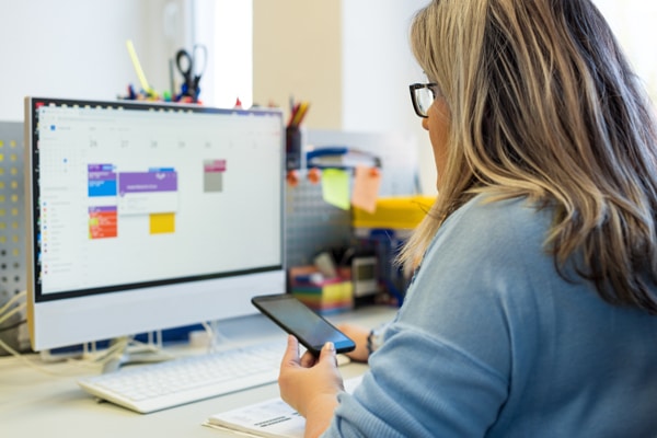 Woman with long blonde hair and glasses sits at a desk, checking a calendar on her computer while holding a smartphone and notepad.