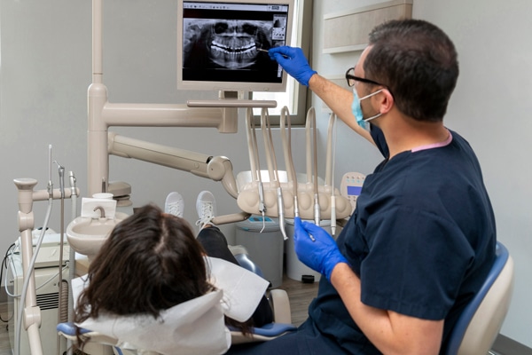 A dentist in gloves and mask points to a dental X-ray on a monitor as a patient reclines, viewing the screen; tools nearby.