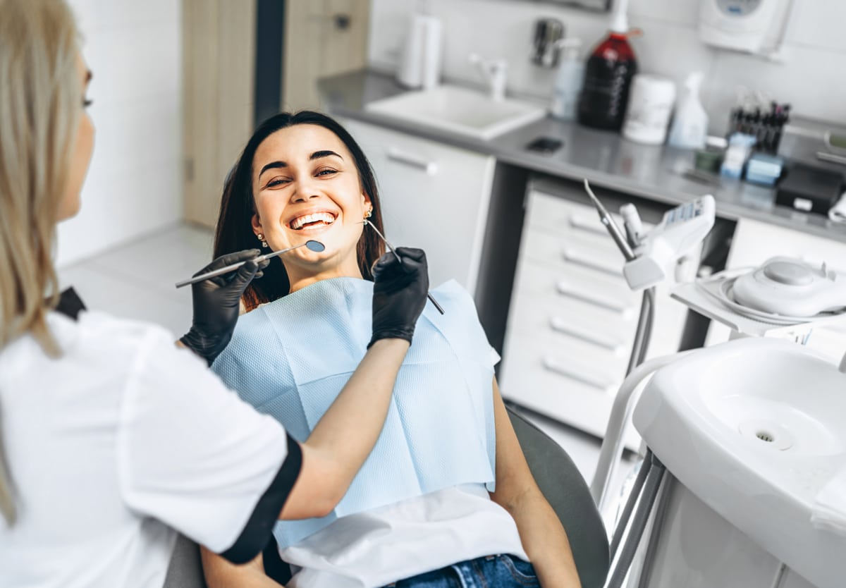 A woman smiles in a dental chair while a gloved dentist examines her teeth with tools in a modern clinic.