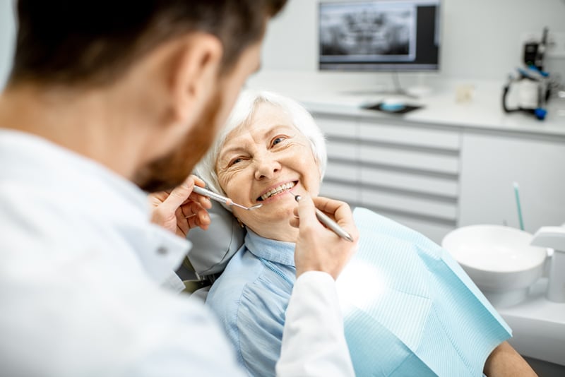 An elderly woman smiles in a dental chair as a dentist examines her teeth; computer and dental equipment are in the background.