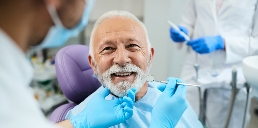 An older man with a white beard smiles in a dental chair as a masked dentist holds tools near his mouth; another staff stands behind.