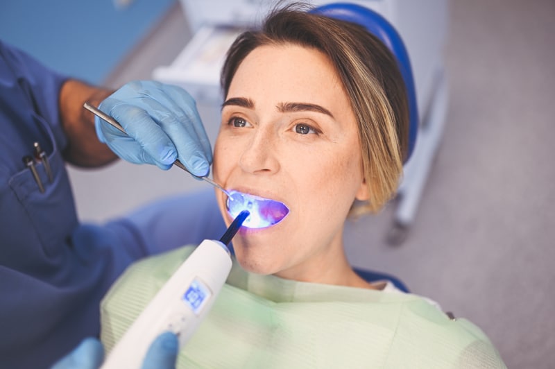 A dentist in blue gloves uses a curing light tool in a woman's open mouth as she sits in a dental chair during treatment.
