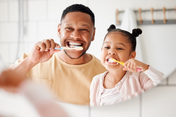 A father and his young daughter happily brush their teeth side by side in front of a bathroom mirror.
