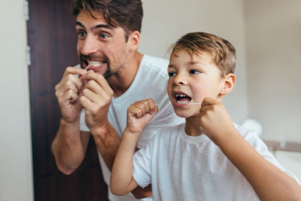 A man and a young boy in white shirts stand together in a bathroom, smiling as they floss their teeth in front of the mirror.