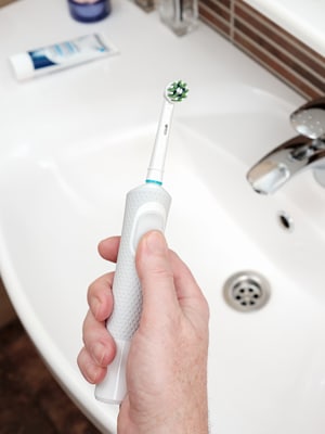 A person’s hand holds an electric toothbrush above a white bathroom sink, with toothpaste on the countertop in the background.