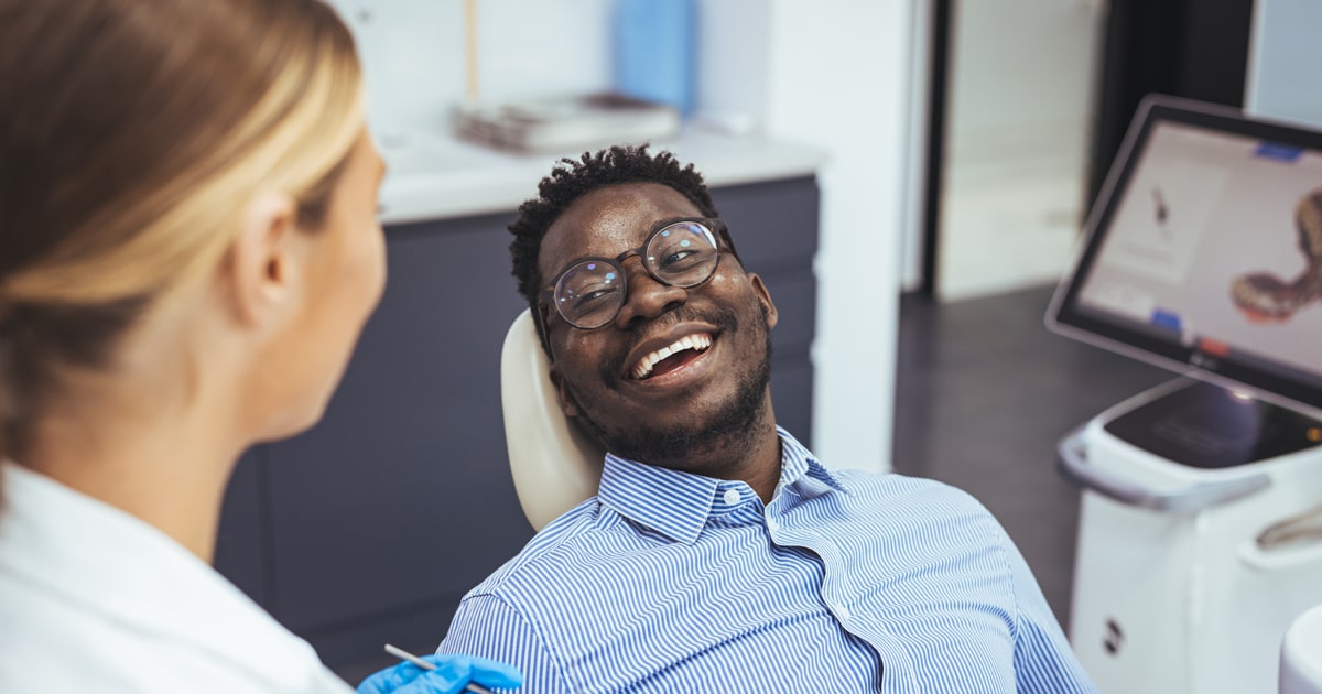 A man with glasses and a striped shirt smiles in a dental chair, facing a dentist. Dental tools and a monitor appear in the background.