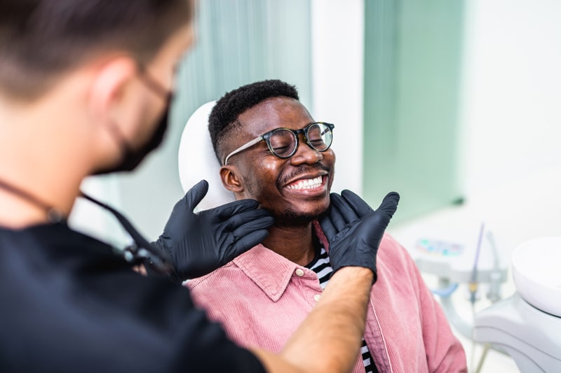 A smiling man with glasses sits in a dental chair as a dentist in black gloves gently examines his jaw and face in a modern clinic.