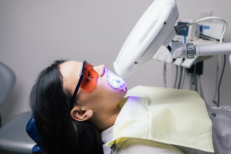 A person in protective glasses reclines in a dental chair as a specialized LED lamp is aimed at their mouth for teeth whitening.