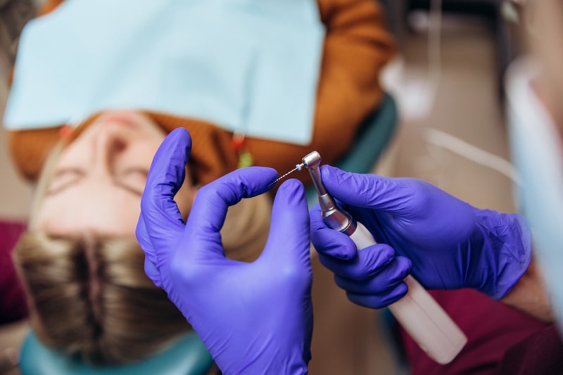 A person lies on a dental chair with a bib as a dentist wearing purple gloves prepares a dental drill, seen from above.