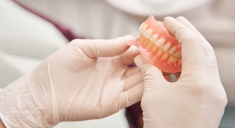 A person in white gloves holds upper and lower dentures, showing artificial teeth and gums in a dental or clinical environment.