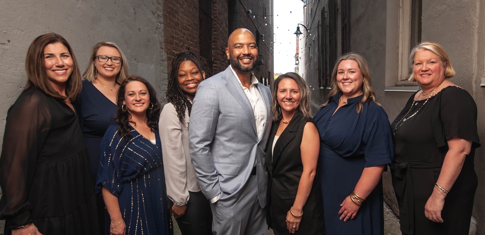 Eight professionally dressed adults, seven women and one man in a blue suit, smile together in an urban brick alleyway.
