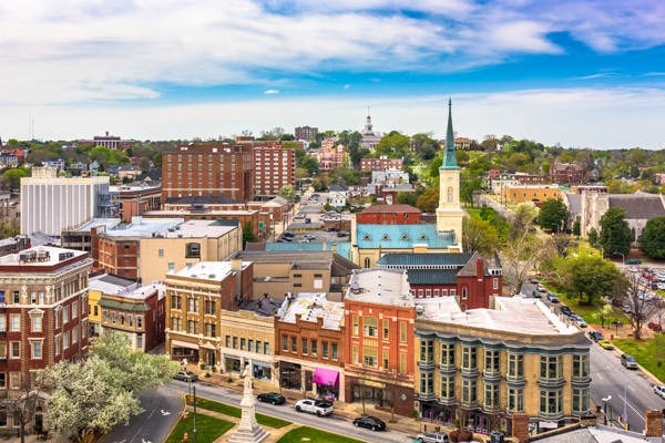 Aerial view of downtown Macon, Georgia with historic buildings, a tall-steepled church, tree-lined streets, and partly cloudy sky.