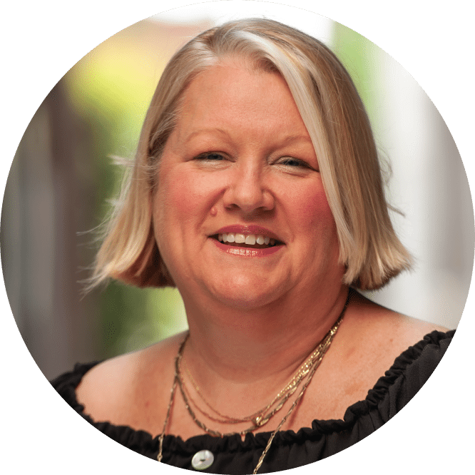 Smiling woman with short blonde hair in a black off-the-shoulder top and gold necklaces, in soft natural light, blurred background.