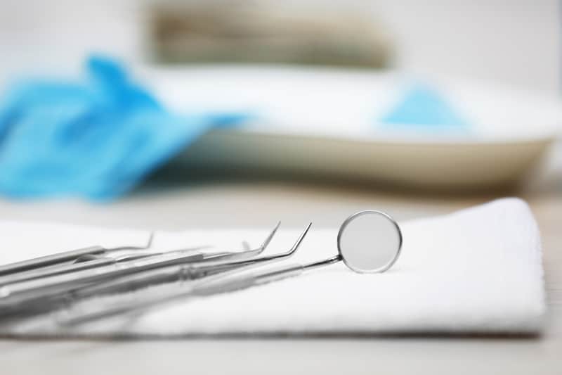 Close-up of dental tools like a mirror and probes on a white cloth, with blue gloves and a tray blurred in the background.