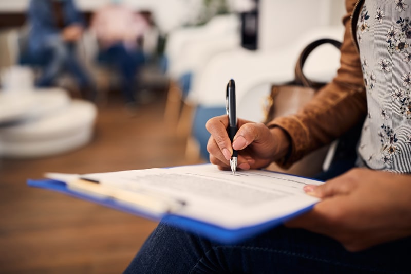 A person in jeans, a brown jacket, and a floral top fills out a clipboard form. A handbag sits close by with a softly blurred background.