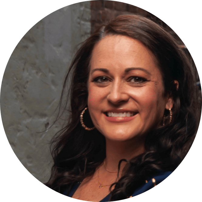 A woman with long dark hair, hoop earrings, and a necklace smiles at the camera in front of a textured gray wall.