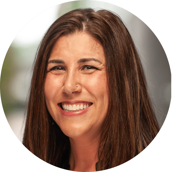 A woman with long brown hair smiles warmly at the camera, a nose piercing visible, standing before a gently blurred background.