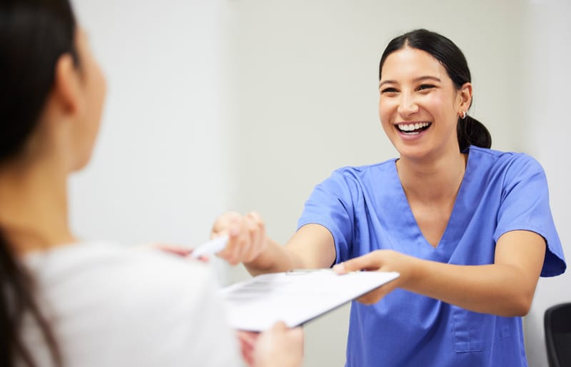 A cheerful healthcare worker in blue scrubs offers a clipboard and pen to a patient across a desk in a well-lit clinic.
