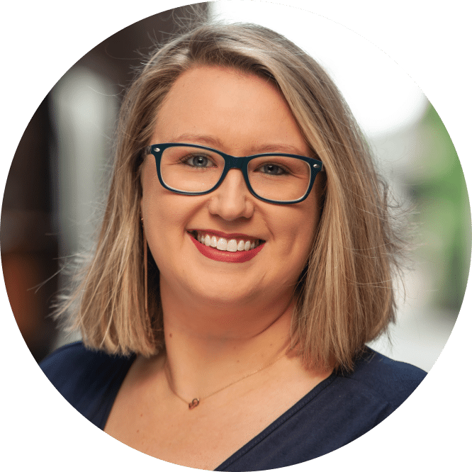 A woman with shoulder-length blonde hair, glasses, a navy top, and a necklace smiles at the camera with a softly blurred background.