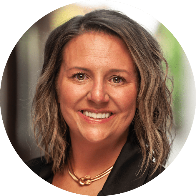 Smiling woman with wavy, shoulder-length brown hair in a black top and gold necklace, standing before a blurred background.