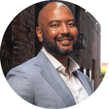 A smiling man with a shaved head and beard, in a light blue suit jacket and white shirt, stands outdoors by brick walls.