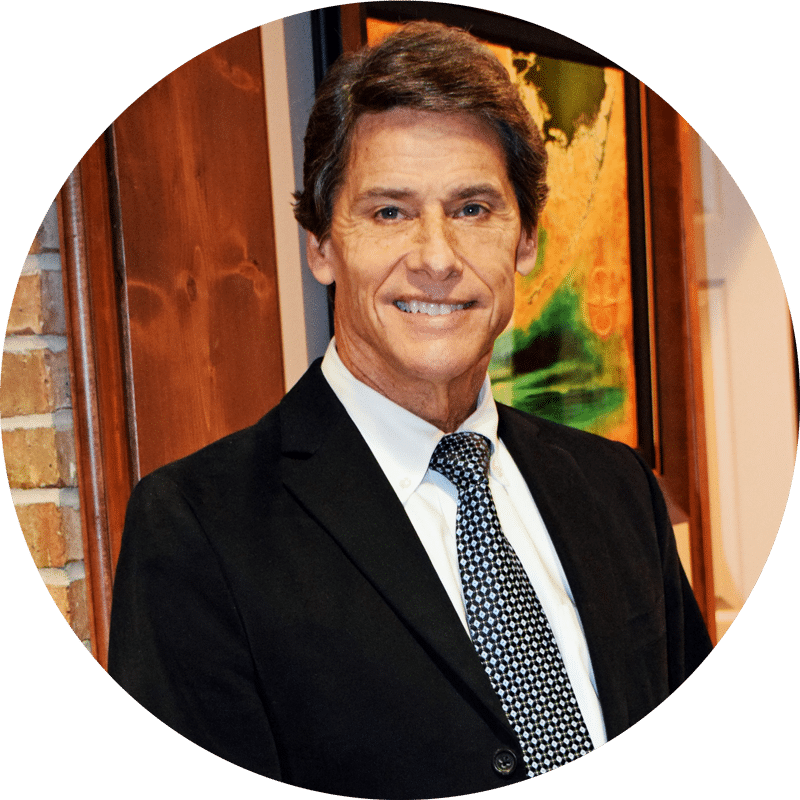 A man with brown hair in a black suit, white shirt, and patterned tie smiles indoors; brick wall and colorful art in the background.