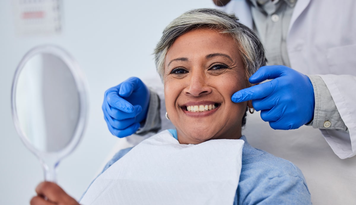 A smiling woman in a dental chair holds a mirror as a dentist in blue gloves points to her cheeks, indicating a dental check-up.